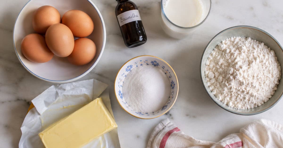 🧈 Bolo Amanteigado da Vovó — Aquele Que o Coração Nunca Esquece 3 Vista de cima dos ingredientes do bolo amanteigado dispostos em bancada de mármore branco: ovos, farinha, manteiga, baunilha, leite e açúcar