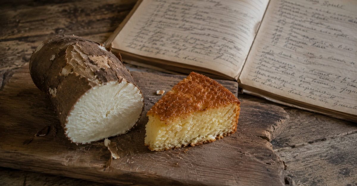 Nunca mais Faça Bolo de Mandioca seco: A Técnica que Muda tudo! 5 Tábua de madeira rústica com mandioca crua com casca ao lado de fatia de bolo assado e caderno de receitas manuscrito ao fundo