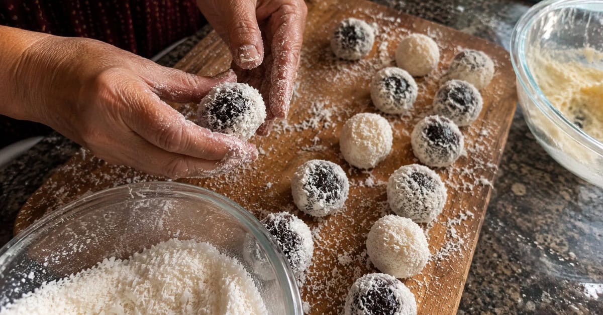 Bolo Olho de Sogra: O Genro vai te Amar — ou será que vai Amar mais o Bolo? 2 Mãos modelando bolinhas de coco e ameixa sobre tábua de madeira enfarinhada para o recheio do Bolo Olho de Sogra