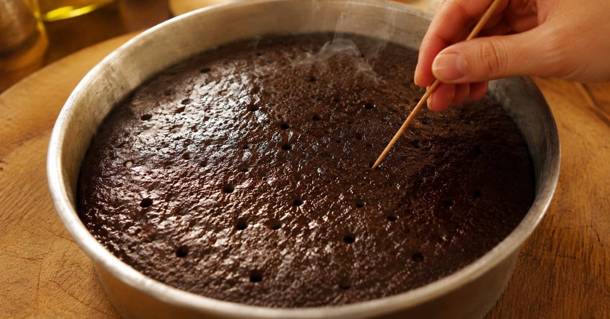 Bolo Nega Maluca sem açúcar: úmido, intenso de chocolate e pronto em menos de 40 minutos 4 Mãos fazendo furos em bolo de chocolate ainda quente na forma para receber a calda da Nega Maluca sem açúcar, com luz dourada natural e bancada rústica ao fundo