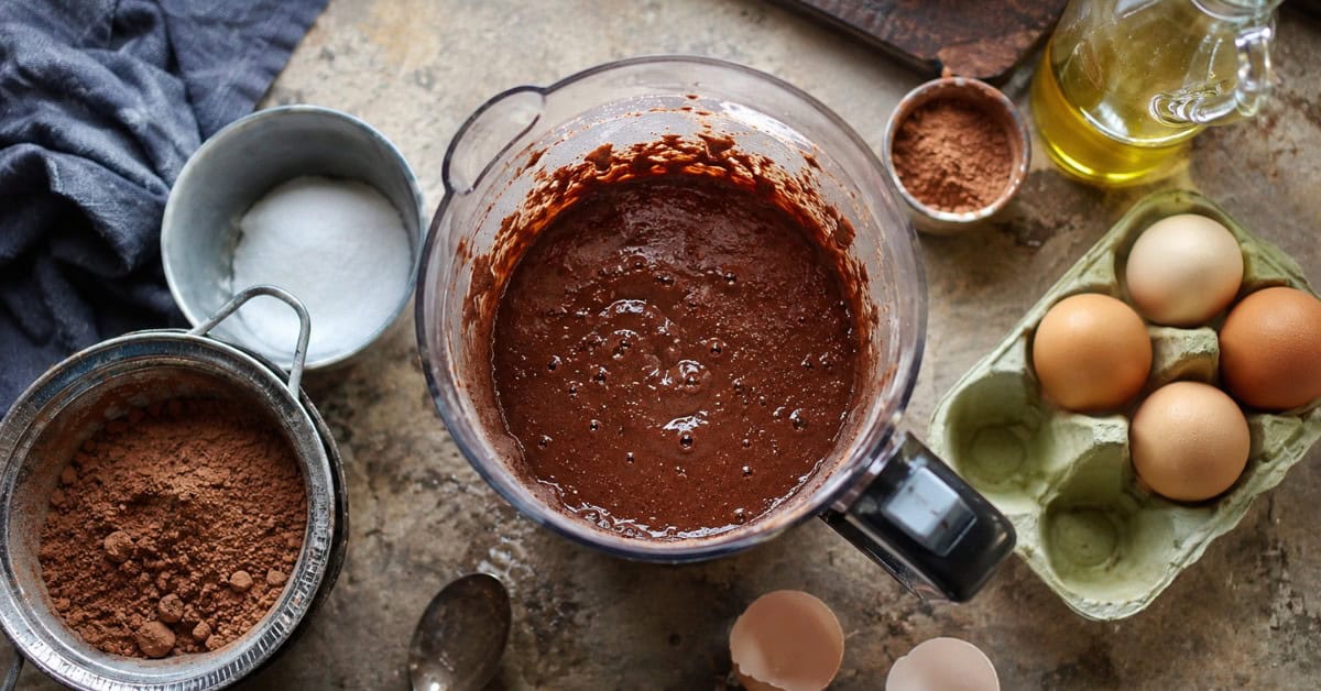 Bolo Nega Maluca sem açúcar: úmido, intenso de chocolate e pronto em menos de 40 minutos 2 Liquidificador com massa de bolo de chocolate cercado por ingredientes como cacau em pó, ovos e adoçante sobre bancada de cozinha
