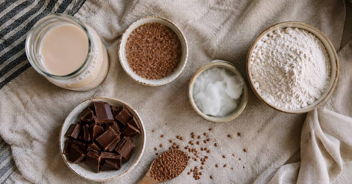 Bolo Nega Maluca sem açúcar: úmido, intenso de chocolate e pronto em menos de 40 minutos 3 Pequenas tigelas com ingredientes substitutos para bolo sem açúcar: leite vegetal, farinha de arroz, óleo de coco, ovo de linhaça e chocolate zero açúcar sobre pano de linho