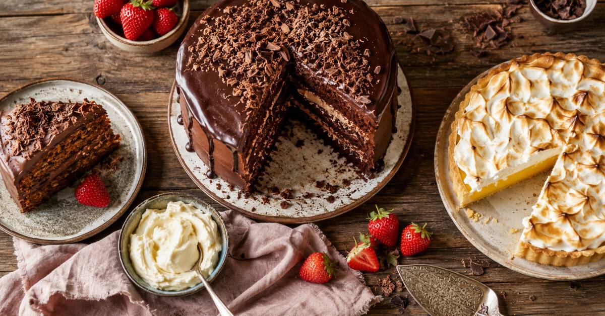 Mesa rústica com bolo de chocolate com ganache escorrendo ao lado de torta de limão com merengue e fatia de bolo de cenoura com cobertura de chocolate