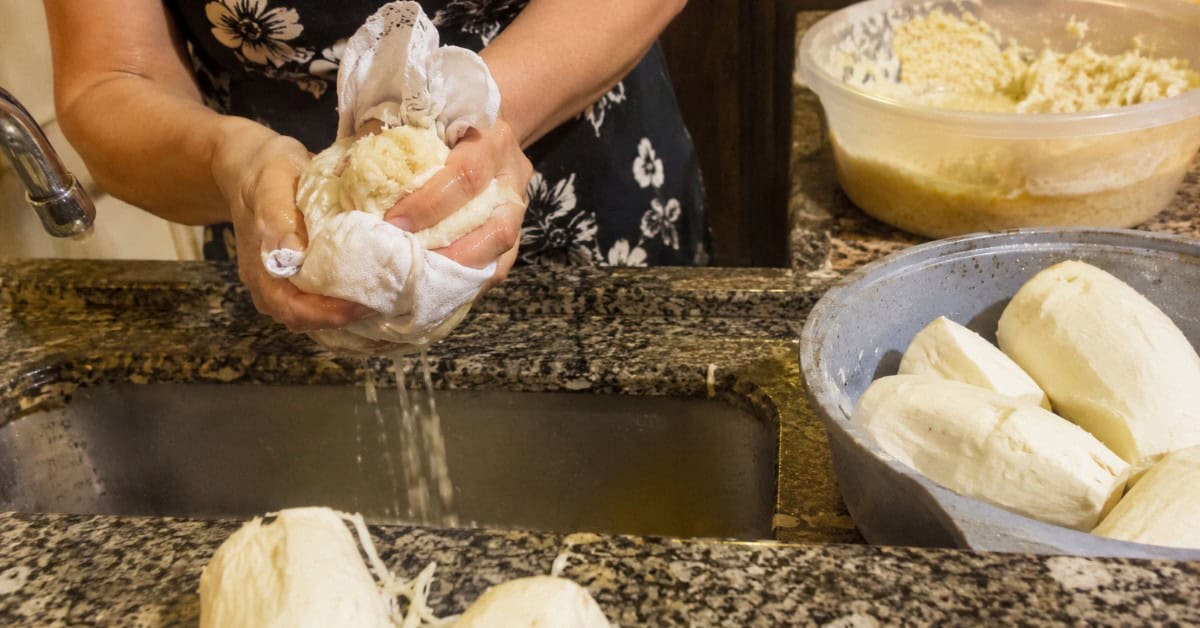 Mãos espremendo mandioca crua ralada em pano de prato branco sobre pia de cozinha doméstica