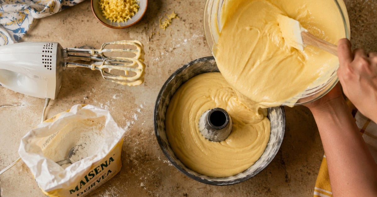 Bolo de Maisena: A Receita Clássica que a Vovó Fazia! 2 Mãos despejando massa de bolo de maisena em forma de burraco untada sobre bancada de cozinha caseira com amido de milho e raspas de limão ao redor