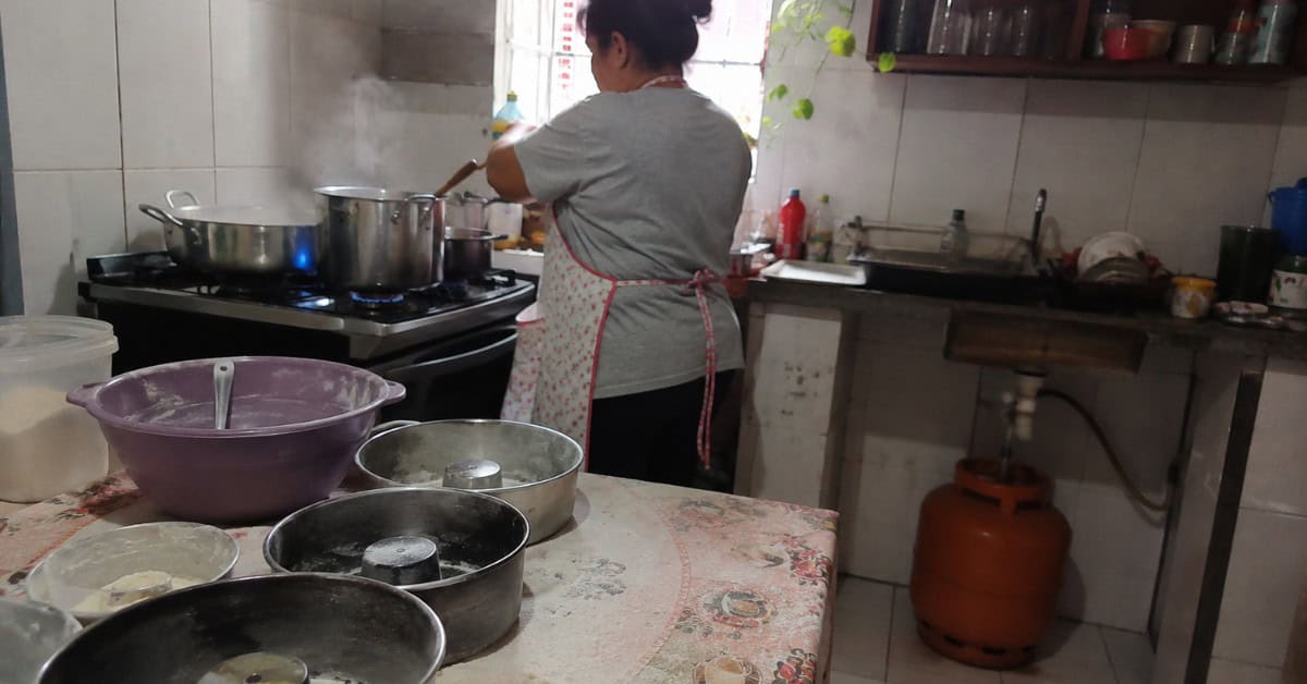 O Gás de Cozinha que Pesava no Bolso — e a Solução que Virou Lei 6 Mulher de avental trabalhando em cozinha doméstica com panelas no fogão aceso, forminhas de bolo na mesa enfarinhada e botijão de gás visível ao fundo