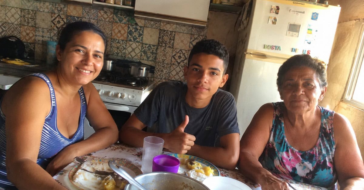 O Gás de Cozinha que Pesava no Bolso — e a Solução que Virou Lei 3 Família simples reunida na cozinha de casa com mãe, filho adolescente e avó à mesa com refeição e fogão a gás ao fundo