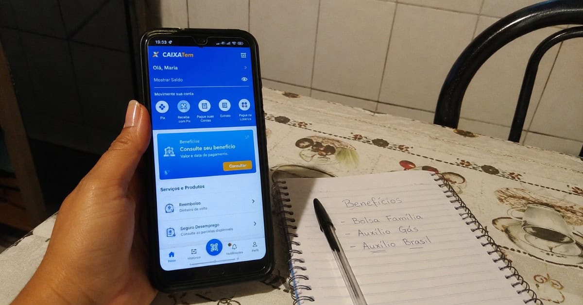 O Gás de Cozinha que Pesava no Bolso — e a Solução que Virou Lei 2 Mãos de mulher segurando celular com aplicativo Caixa Tem aberto sobre mesa de cozinha com caderno de anotações de benefícios sociais