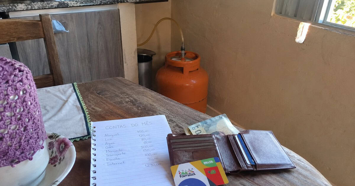 Auxílio Gás (Gás do Povo) - Botijão de gás laranja de 13kg sobre mesa de cozinha simples ao lado de carteira aberta com cartão do Bolsa Família e anotações de contas do mês