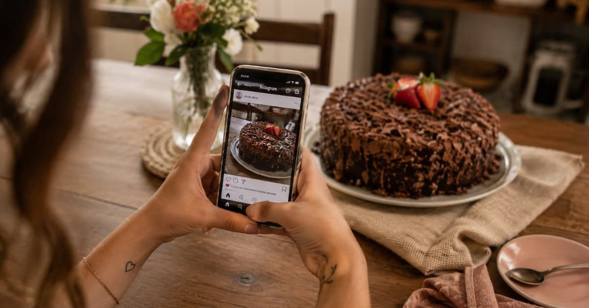 Como Fazer 5 Mil Reais Vendendo Bolo: O Guia Honesto para Transformar Sua Cozinha em Negócio 5 Mãos de mulher segurando celular fotografando bolo de chocolate caseiro sobre mesa com flores e tecido de linho, com Instagram aberto na tela