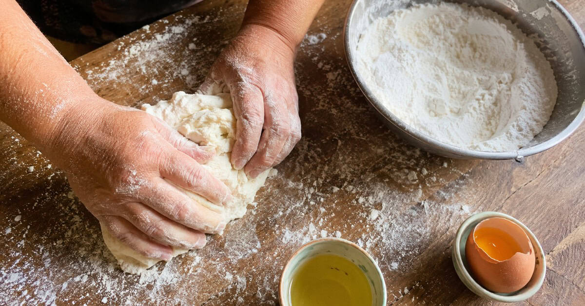 Mãos sovando a massa de biscoito de polvilho em superfície de madeira enfarinhada com ingredientes ao redor