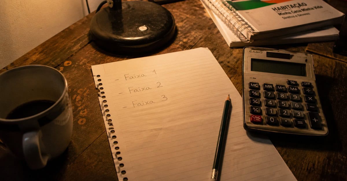 Como Conseguir uma Casa na Minha Casa Minha Vida 2 Papel com anotações à mão das faixas de renda do programa Minha Casa Minha Vida 2026 ao lado de uma calculadora e uma xícara de café