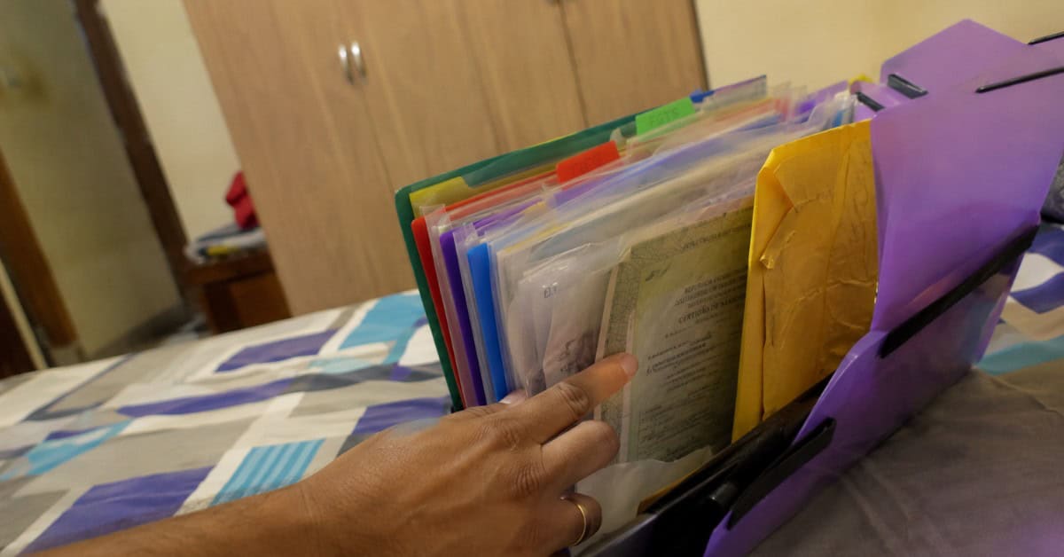 Como Conseguir uma Casa na Minha Casa Minha Vida 5 Pasta sanfonada com documentos para financiamento habitacional aberta sobre cama, com mão masculina apontando para os papéis do Minha Casa Minha Vida